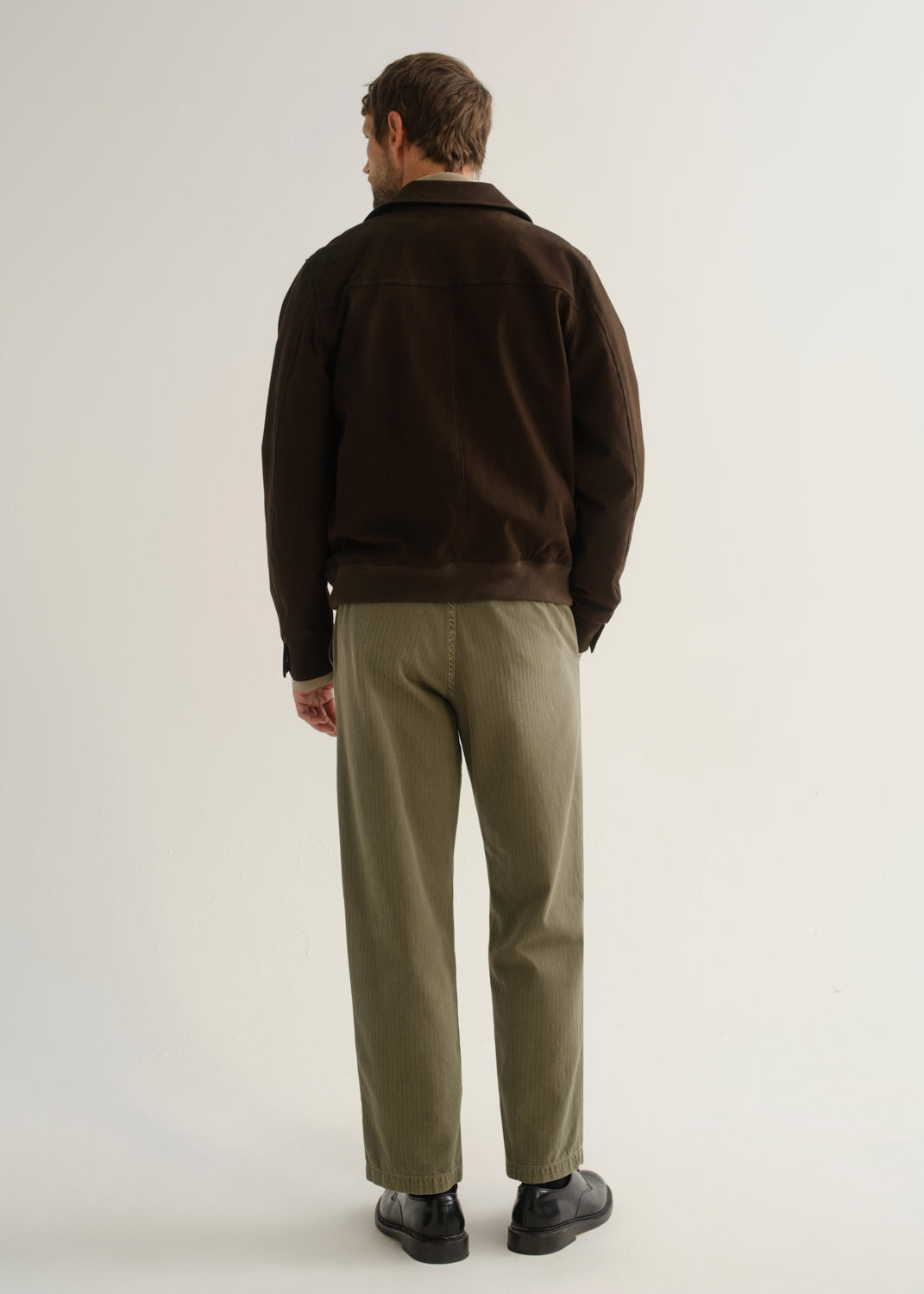 Person wearing olive green relaxed straight leg pants with a dark brown jacket and black boots, back full body view on a light grey background.