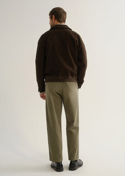 Person wearing olive green relaxed straight leg pants with a dark brown jacket and black boots, back full body view on a light grey background.
