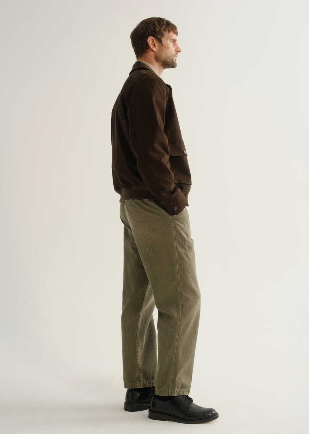 Person wearing olive green relaxed straight leg pants with a dark brown jacket and black boots, side full body view on a light grey background.