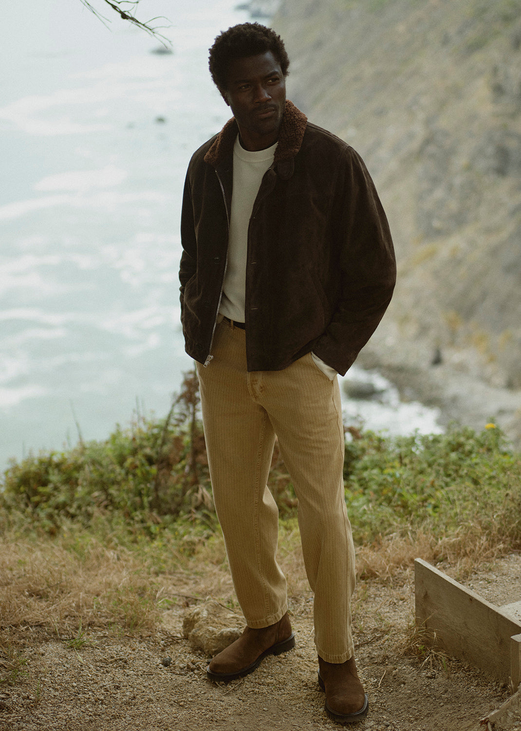 A man stands outdoors by the ocean wearing a dark brown suede jacket, tan pants, and brown boots.
