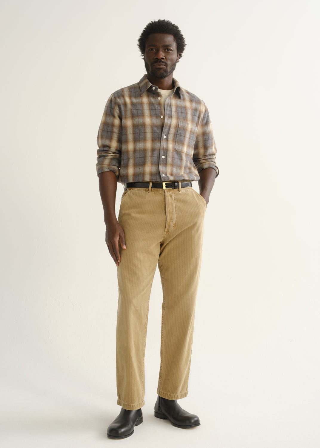 Person wearing tan straight leg corduroy pants with a multicolor plaid button-up shirt and black shoes, front full body view on a light grey background.