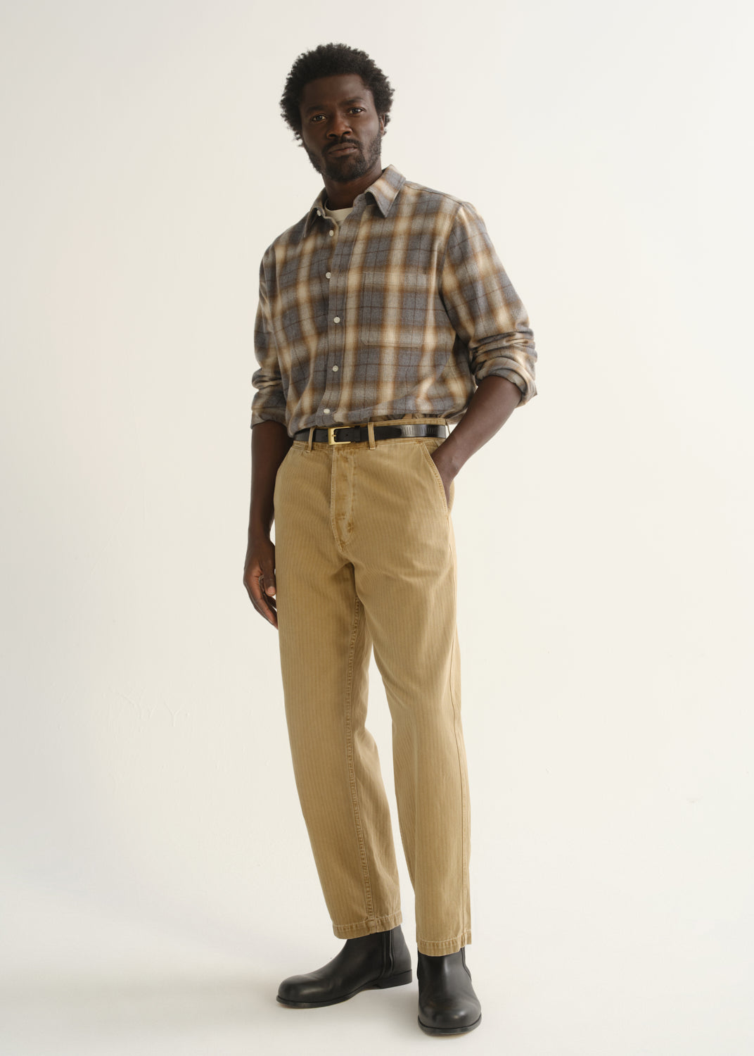 Person wearing tan straight leg corduroy pants with a multicolor plaid button-up shirt and black shoes, angled front full body view on a light grey background.