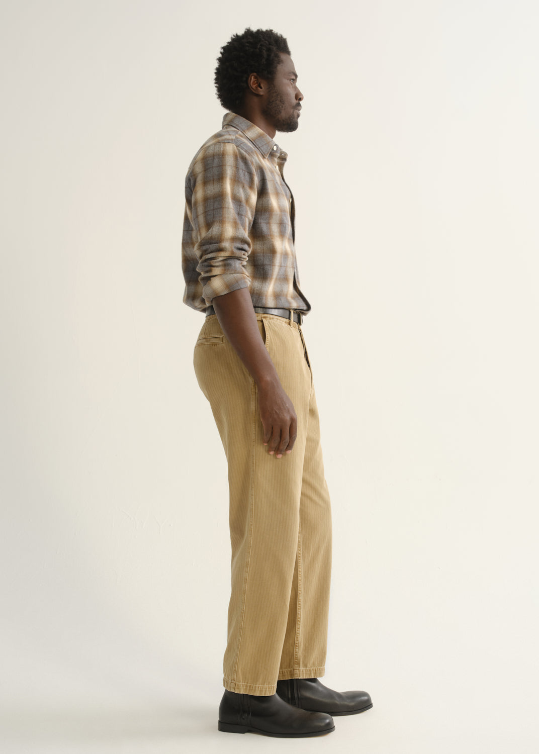 Person wearing tan straight leg corduroy pants with a multicolor plaid button-up shirt and black shoes, side full body view on a light grey background.