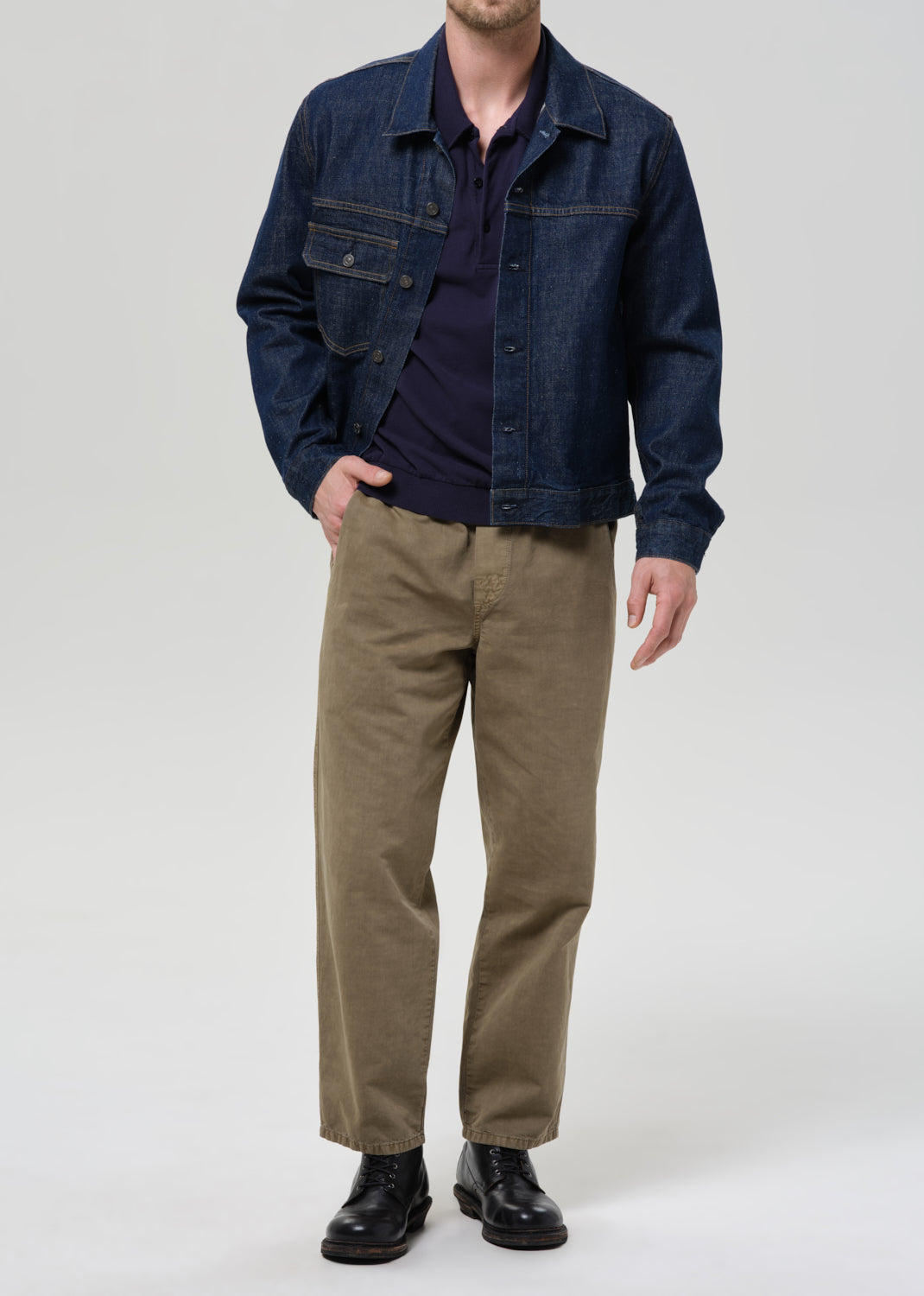 Person wearing a dark denim jacket, navy shirt, and beige pants on a plain background