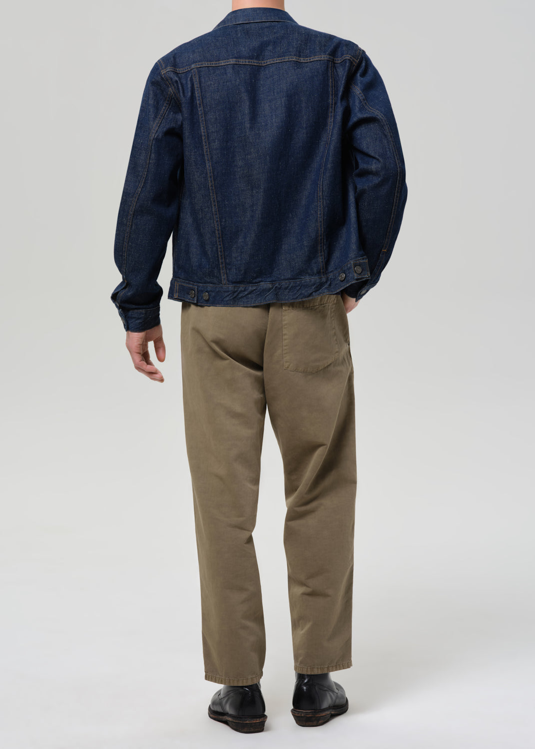 Person wearing a dark denim jacket, navy shirt, and beige pants on a plain background