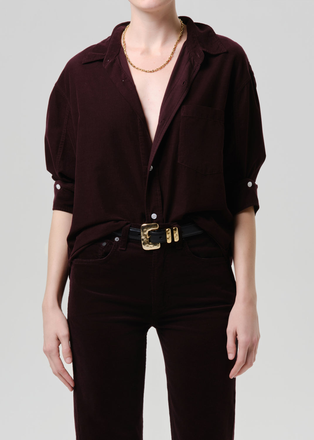 Person wearing a dark purple shirt with short sleeves and a gold belt on a grey background