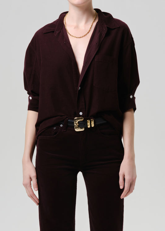 Person wearing a dark purple shirt with short sleeves and a gold belt on a grey background