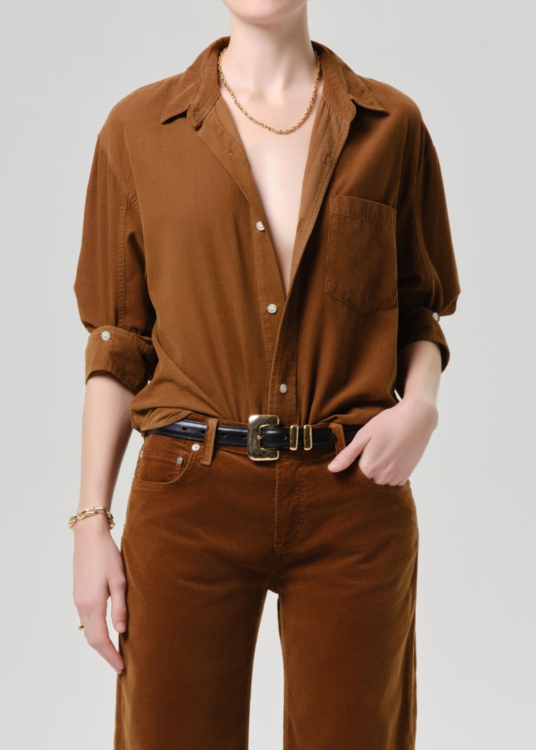 Person wearing a brown shirt and pants with a necklace on a grey background