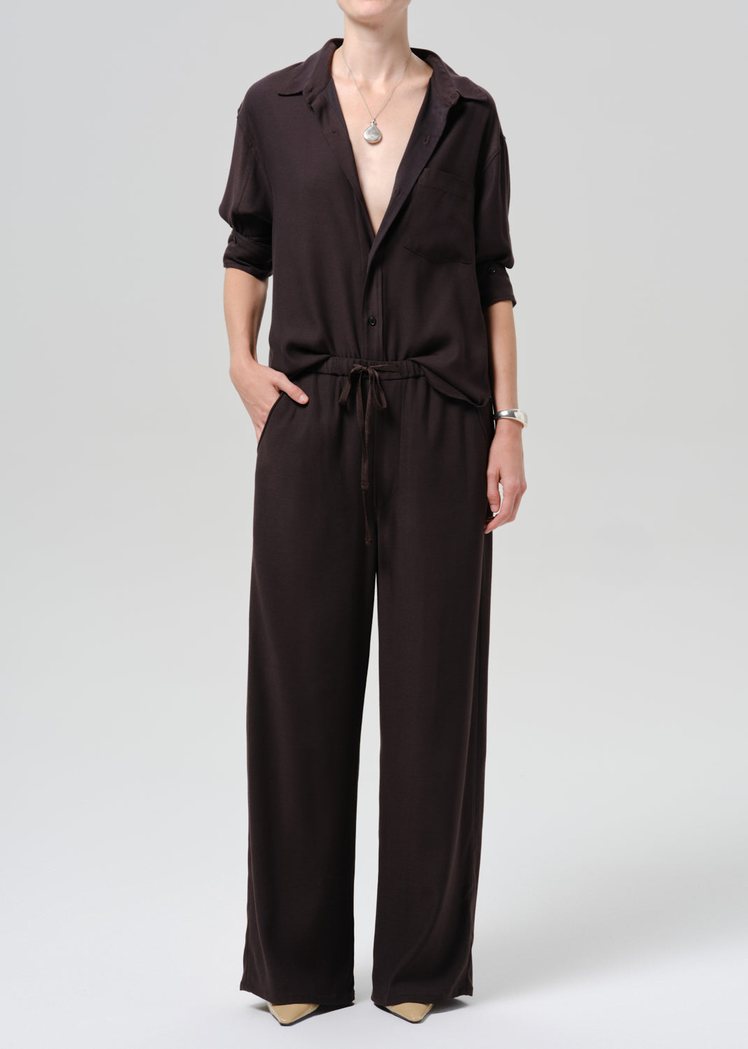 A model wearing a dark brown collared jumpsuit with short sleeves, wide legs, and a matching waist tie, photographed against a grey background.