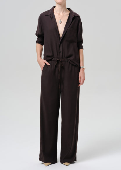 A model wearing a dark brown collared jumpsuit with short sleeves, wide legs, and a matching waist tie, photographed against a grey background.