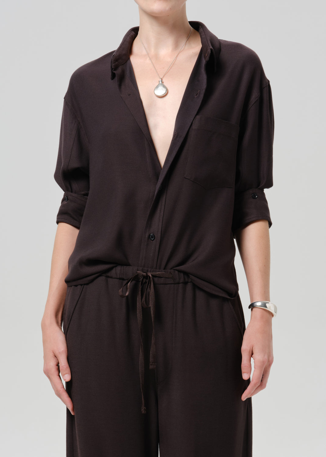 A model wearing a dark brown, long-sleeved collared jumpsuit with a tie waist and matching pants, styled with a silver pendant necklace and a silver bracelet against a grey background.