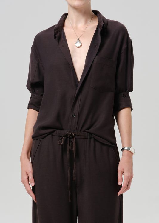 A model wearing a dark brown, long-sleeved collared jumpsuit with a tie waist and matching pants, styled with a silver pendant necklace and a silver bracelet against a grey background.