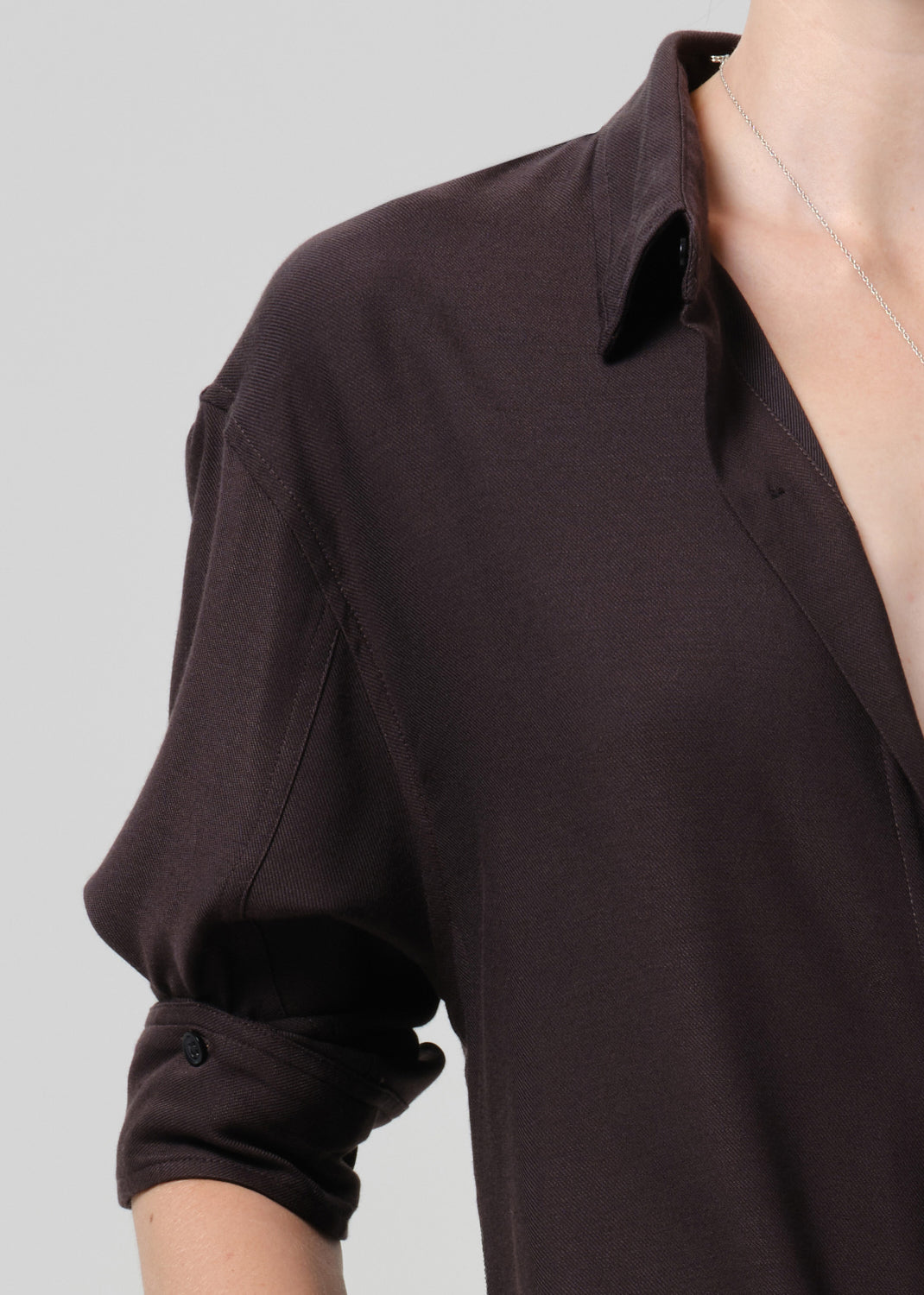 Close-up of a person wearing a dark brown button-up shirt with rolled sleeves, shown against a grey background.