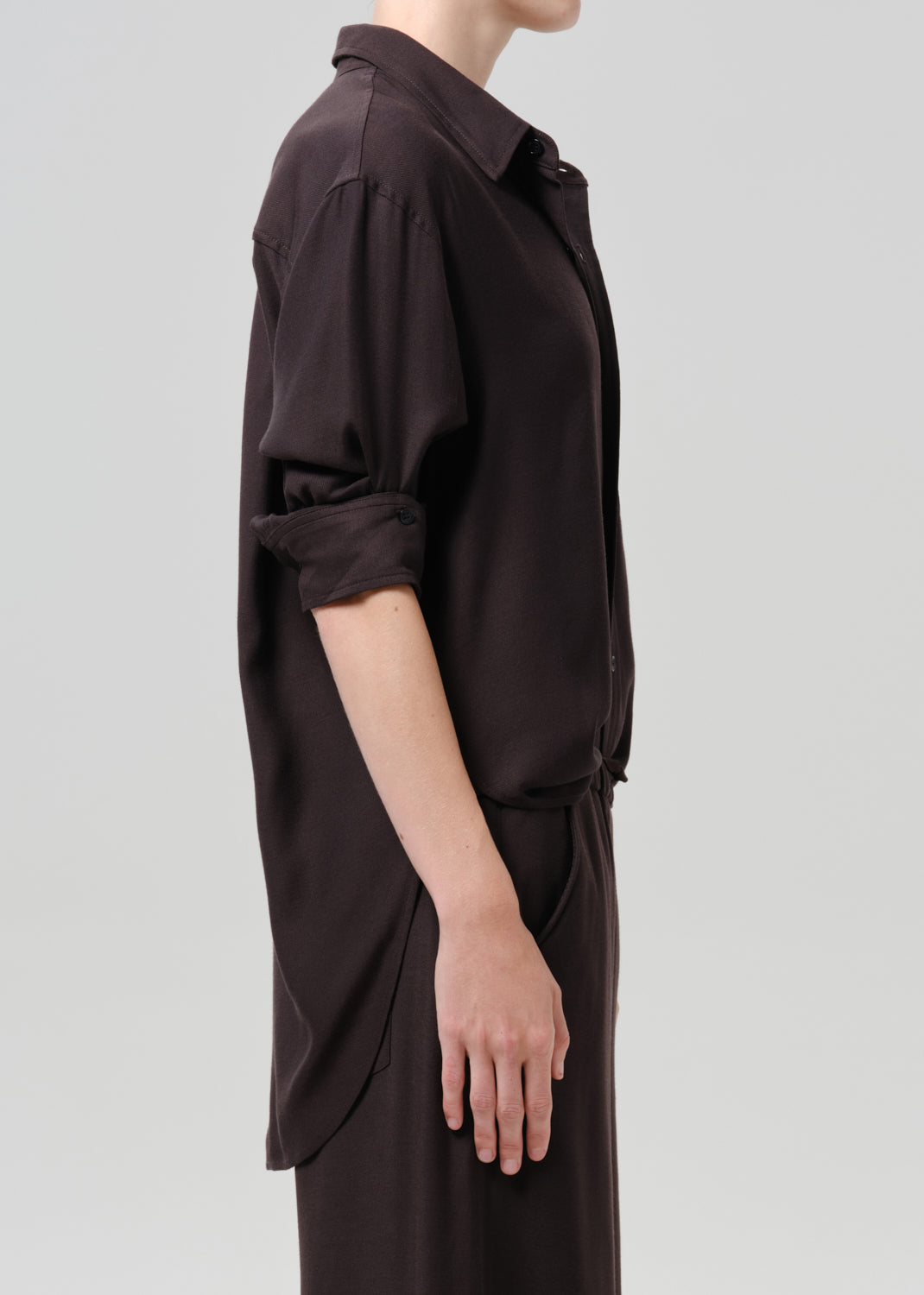 Side profile of a model wearing a dark brown long-sleeve collared shirt dress with rolled-up sleeves, shown against a grey background.