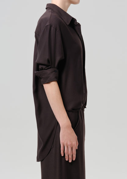 Side profile of a model wearing a dark brown long-sleeve collared shirt dress with rolled-up sleeves, shown against a grey background.