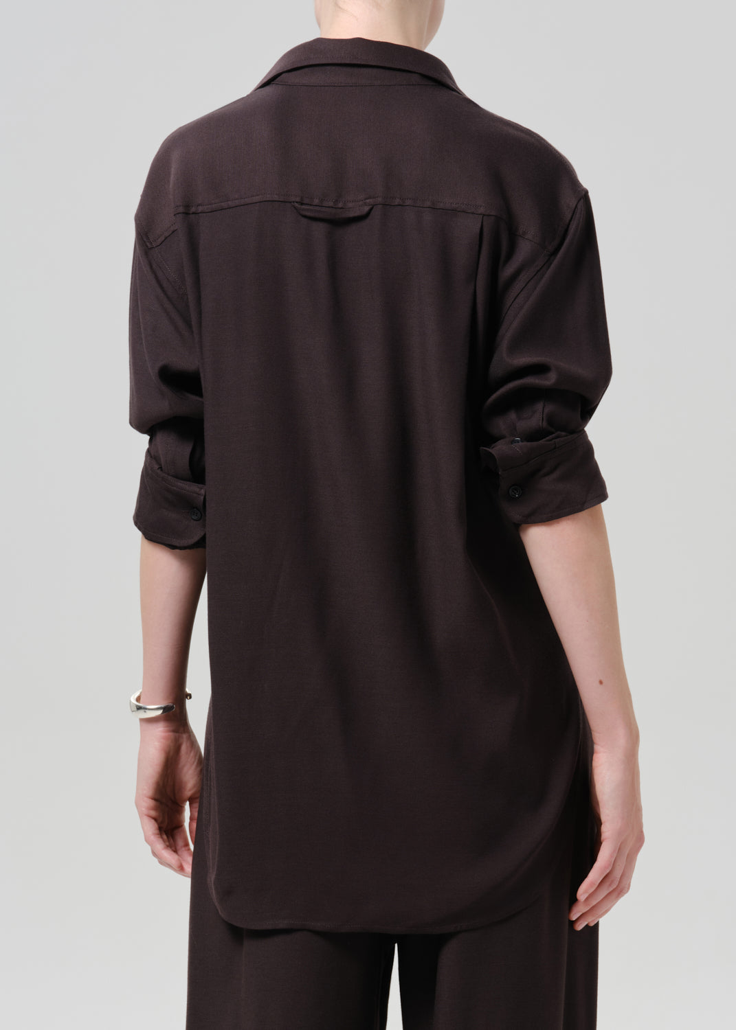 A person shown from the back wearing an oversized dark brown button-up shirt with rolled sleeves and matching dark pants, photographed against a grey background.