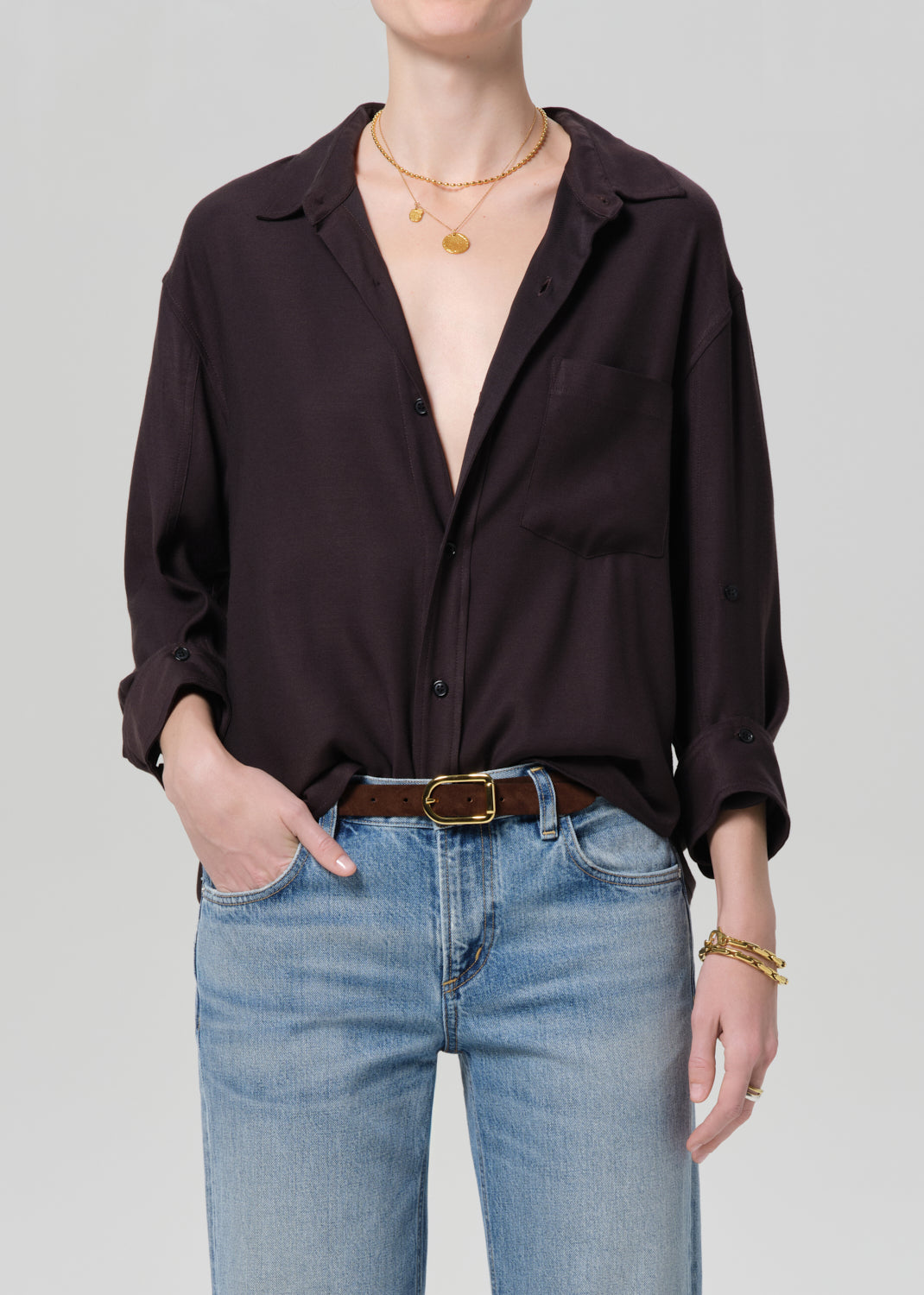 Person wearing a brown shirt and blue jeans with gold jewelry on a plain background