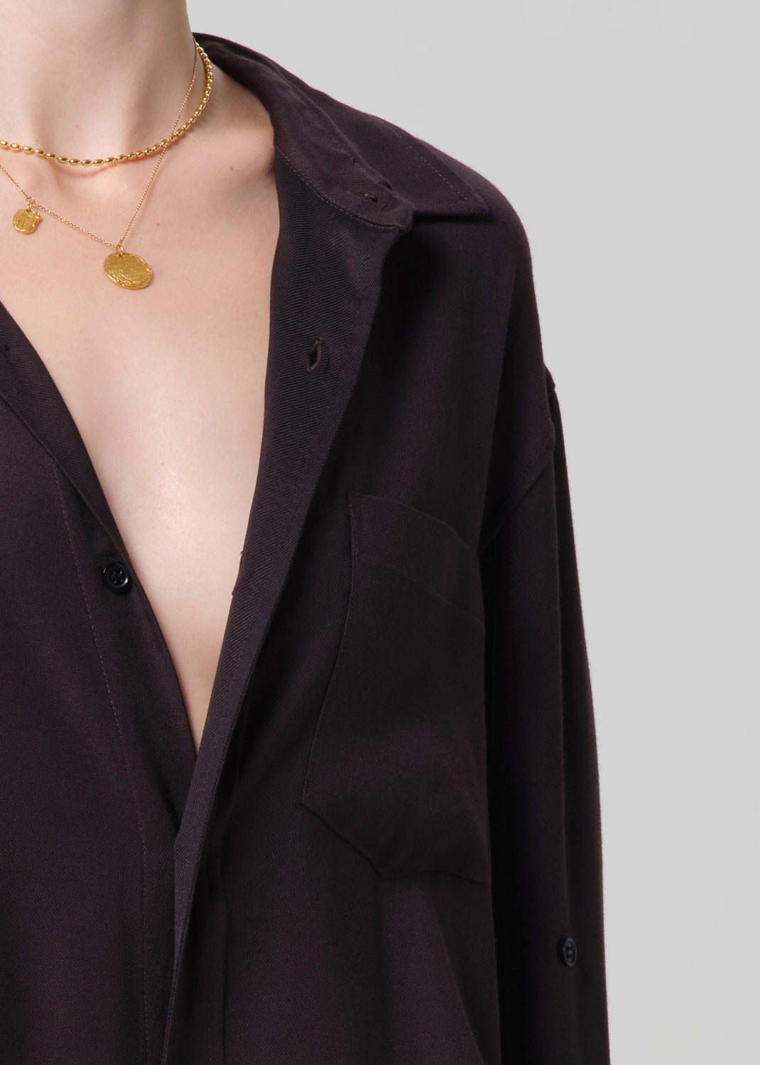 Close-up of a person wearing a dark brown shirt with a deep neckline, gold necklaces, and a plain background.