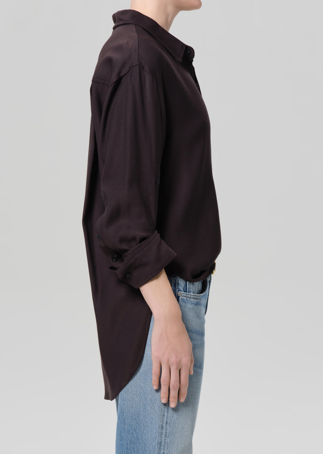 Person wearing a brown shirt and blue jeans with gold jewelry on a plain background
