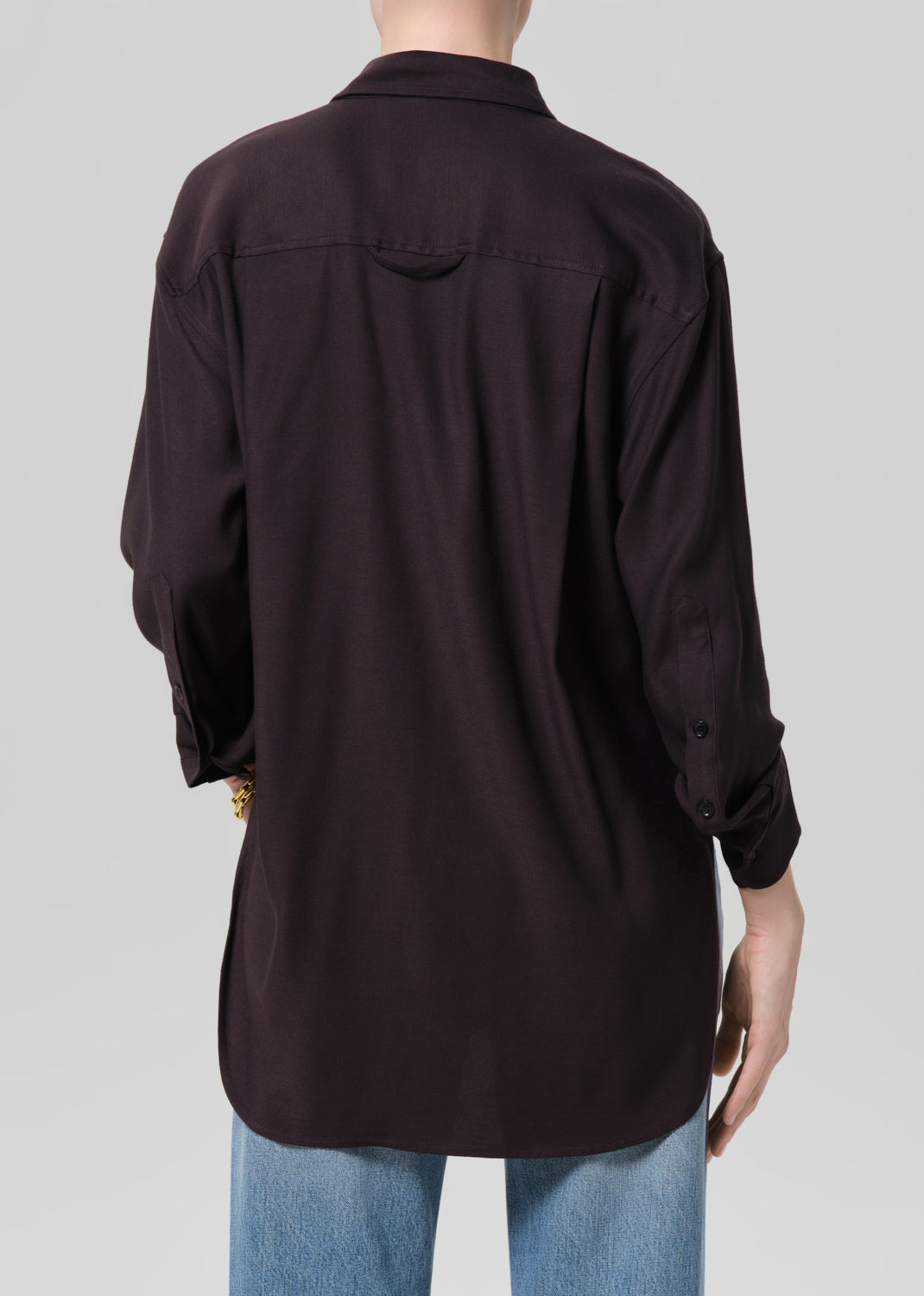 Person wearing a brown shirt and blue jeans with gold jewelry on a plain background