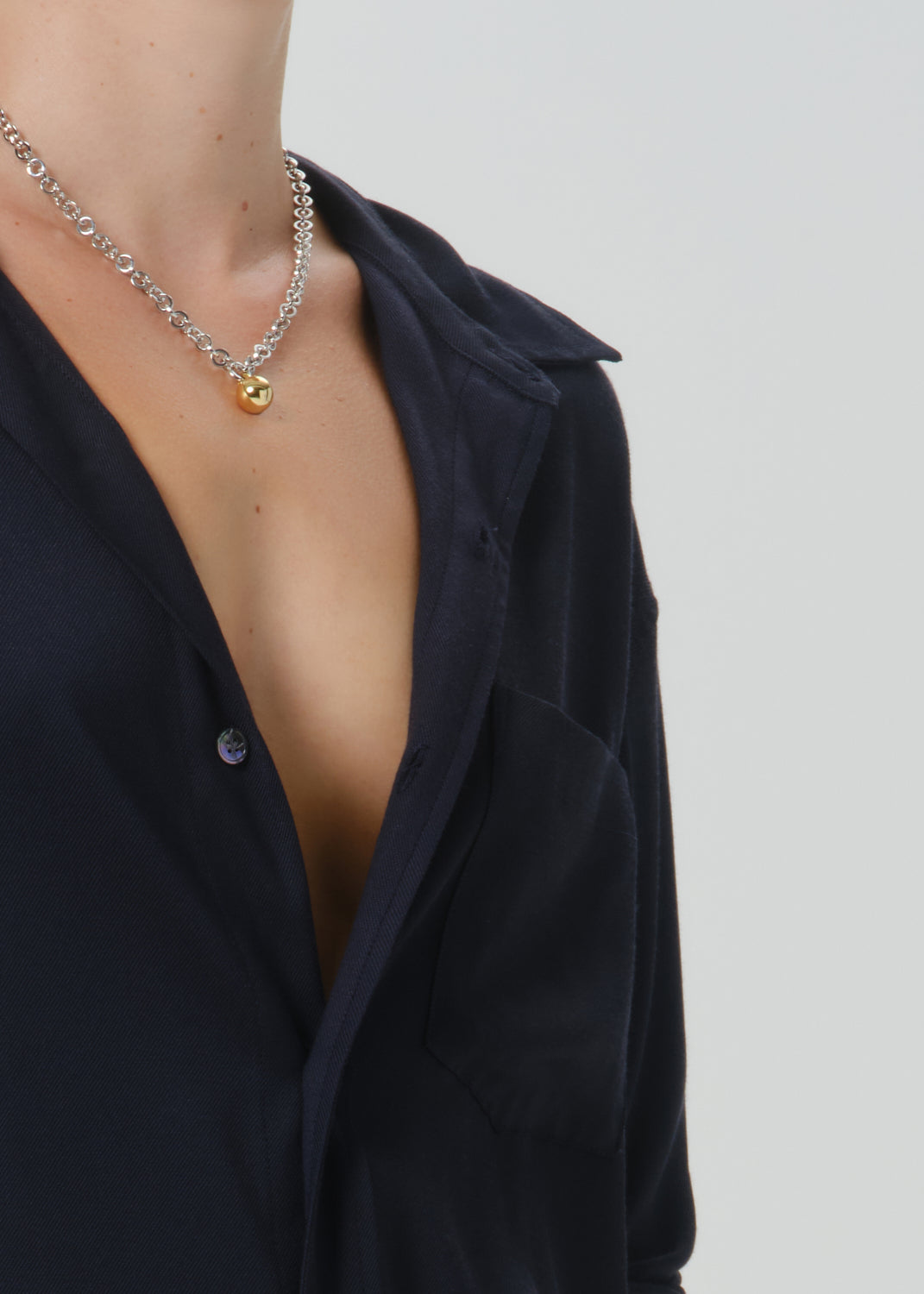 A close-up of a model wearing a silver chain necklace with a small gold pendant, styled with a dark navy button-down shirt against a grey background.