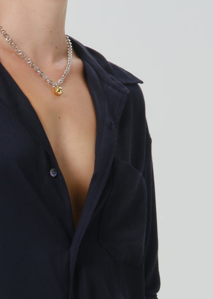 A close-up of a model wearing a silver chain necklace with a small gold pendant, styled with a dark navy button-down shirt against a grey background.