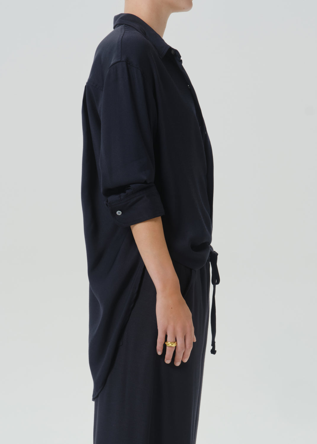 A side-profile view of a person wearing a dark navy long-sleeve shirt dress with rolled-up sleeves and a drawstring tie at the waist, paired with matching dark pants, against a grey background.