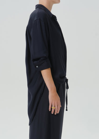 A side-profile view of a person wearing a dark navy long-sleeve shirt dress with rolled-up sleeves and a drawstring tie at the waist, paired with matching dark pants, against a grey background.