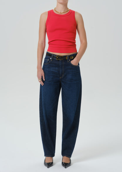 A model wearing a bright red ribbed sleeveless crop top and dark blue wide-leg jeans with a black belt, standing against a grey background.