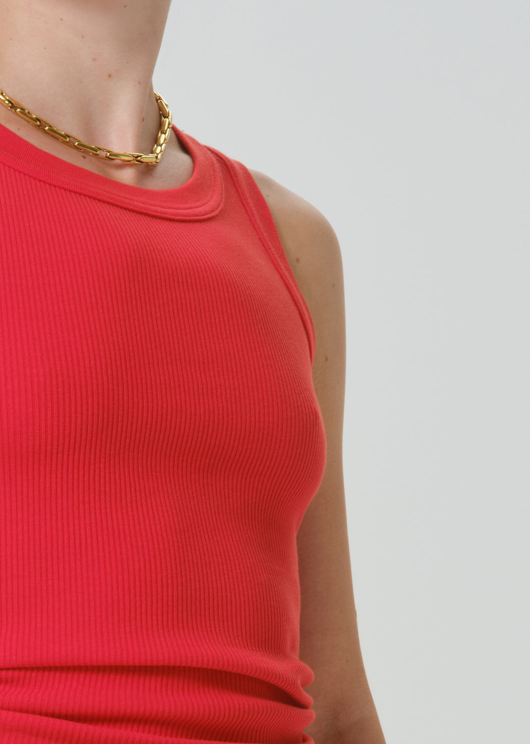 Close-up of a person wearing a bright coral-red ribbed sleeveless top and a chunky gold chain necklace against a grey background.