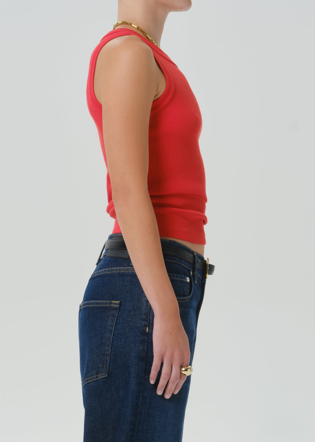 Side profile of a model wearing a bright red ribbed knit tank top and dark blue jeans against a grey background.
