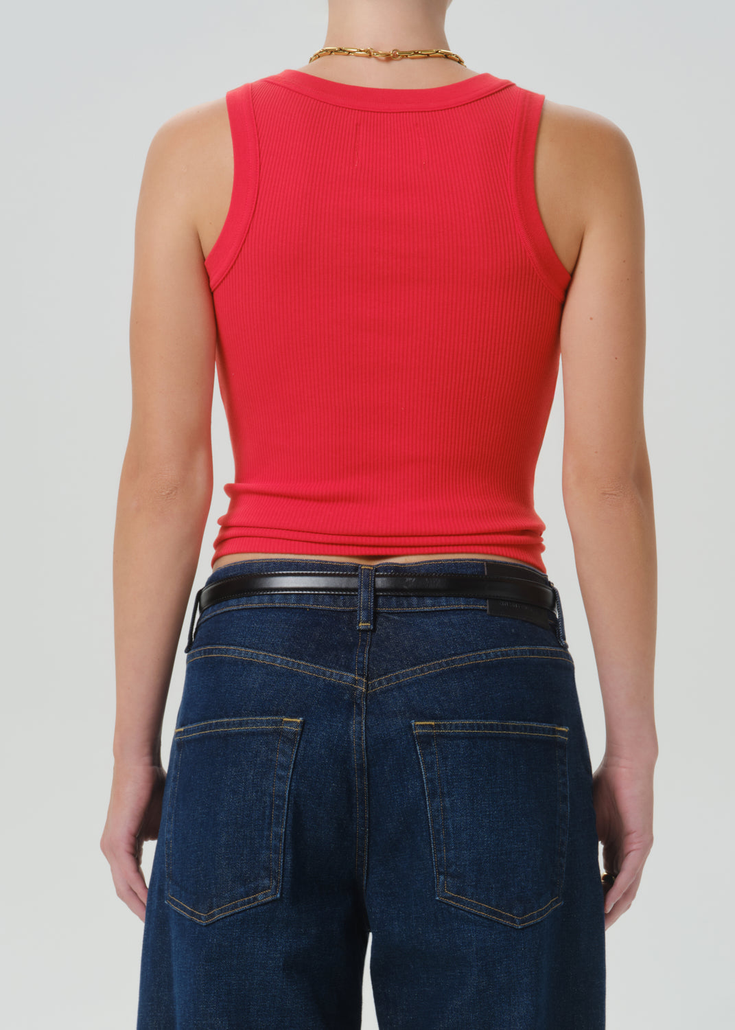 A person shown from the back wearing a bright red ribbed sleeveless tank top, dark blue jeans, and a black belt against a grey background.