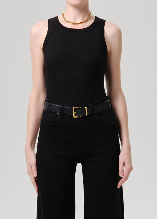 A model wearing a black sleeveless top, black high-waisted pants, a black belt with a gold buckle, and a gold chain necklace, shot against a grey background.