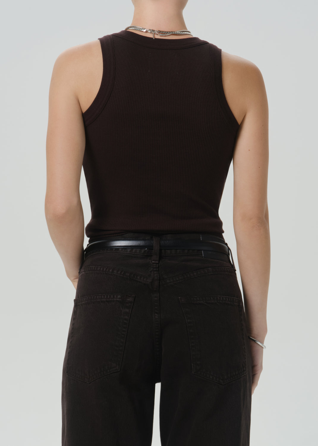 Rear view of a model wearing a dark brown ribbed sleeveless tank top and black jeans against a grey background.