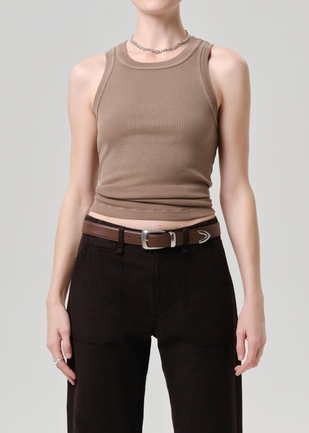 A model wearing a ribbed sleeveless crop top in taupe brown, paired with black pants and a brown belt, against a grey background.