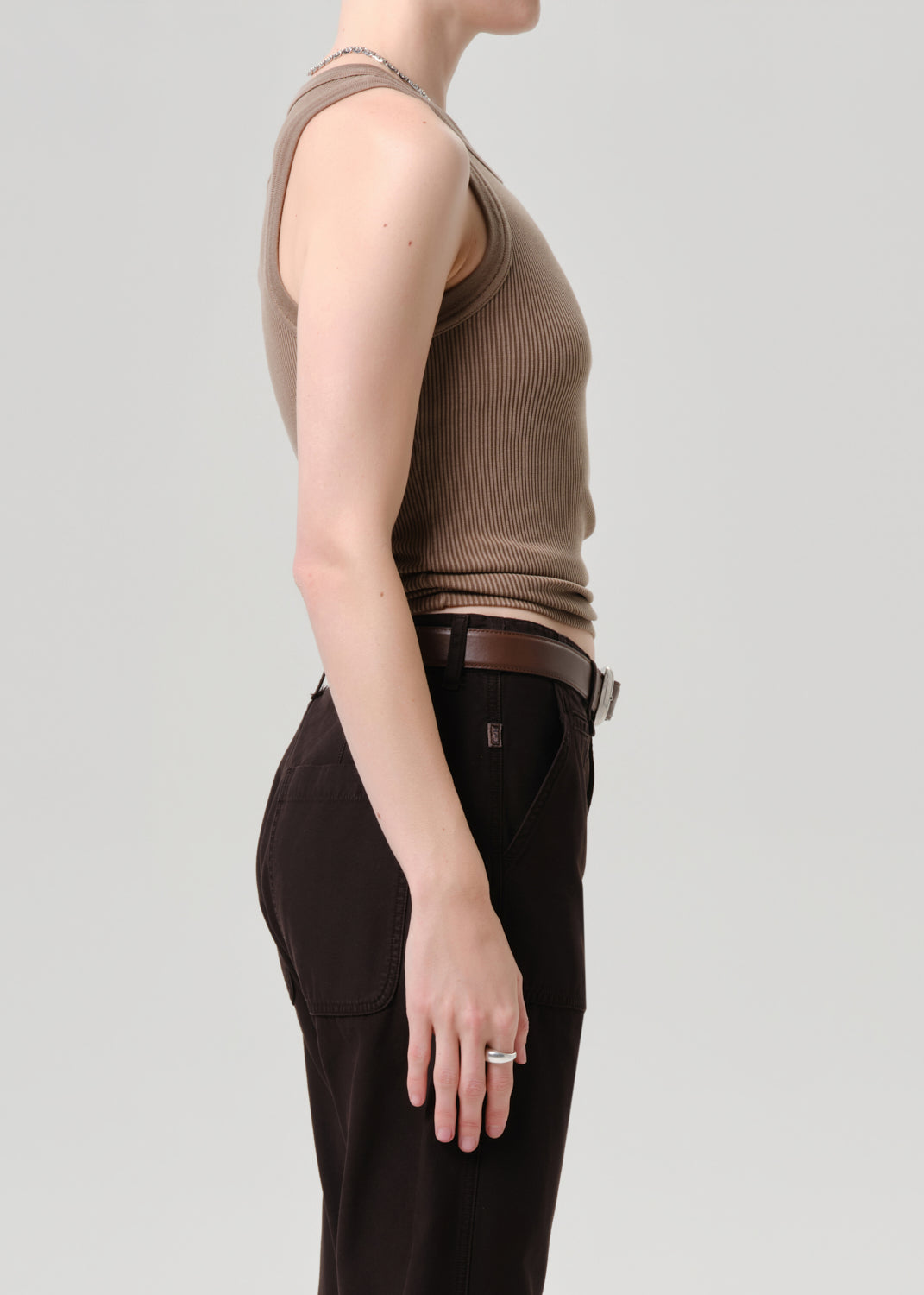 Side profile of a model wearing a ribbed sleeveless knit top in taupe brown and dark brown trousers with a brown belt, against a grey background.