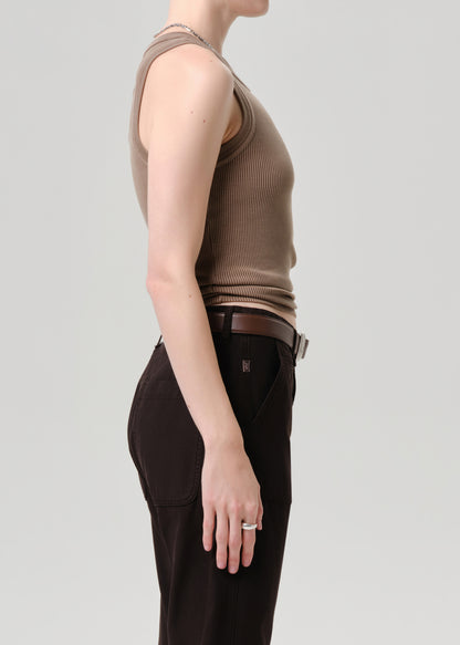 Side profile of a model wearing a ribbed sleeveless knit top in taupe brown and dark brown trousers with a brown belt, against a grey background.