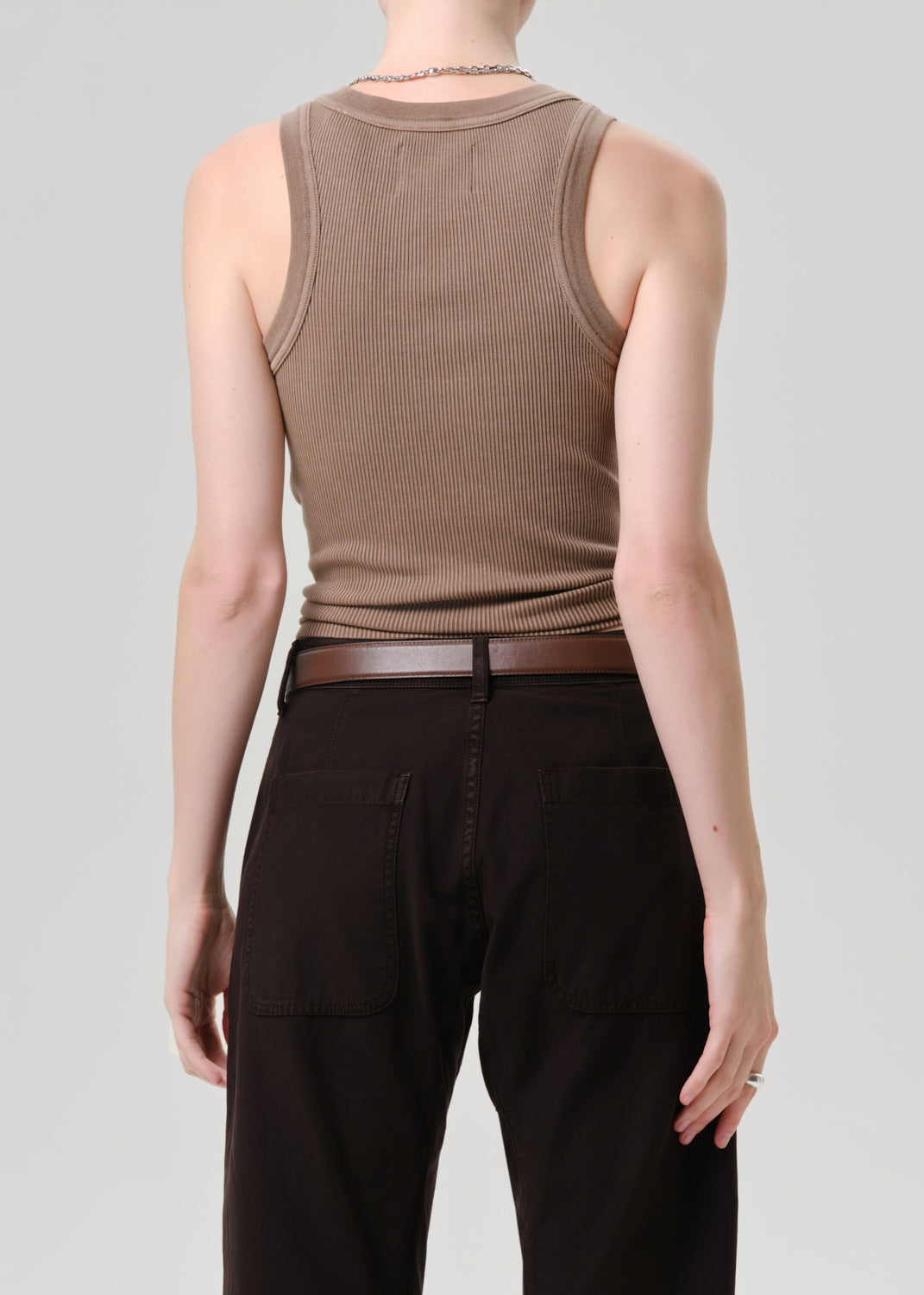 Rear view of a model wearing a ribbed brown tank top and dark brown pants with a matching belt, set against a grey background.
