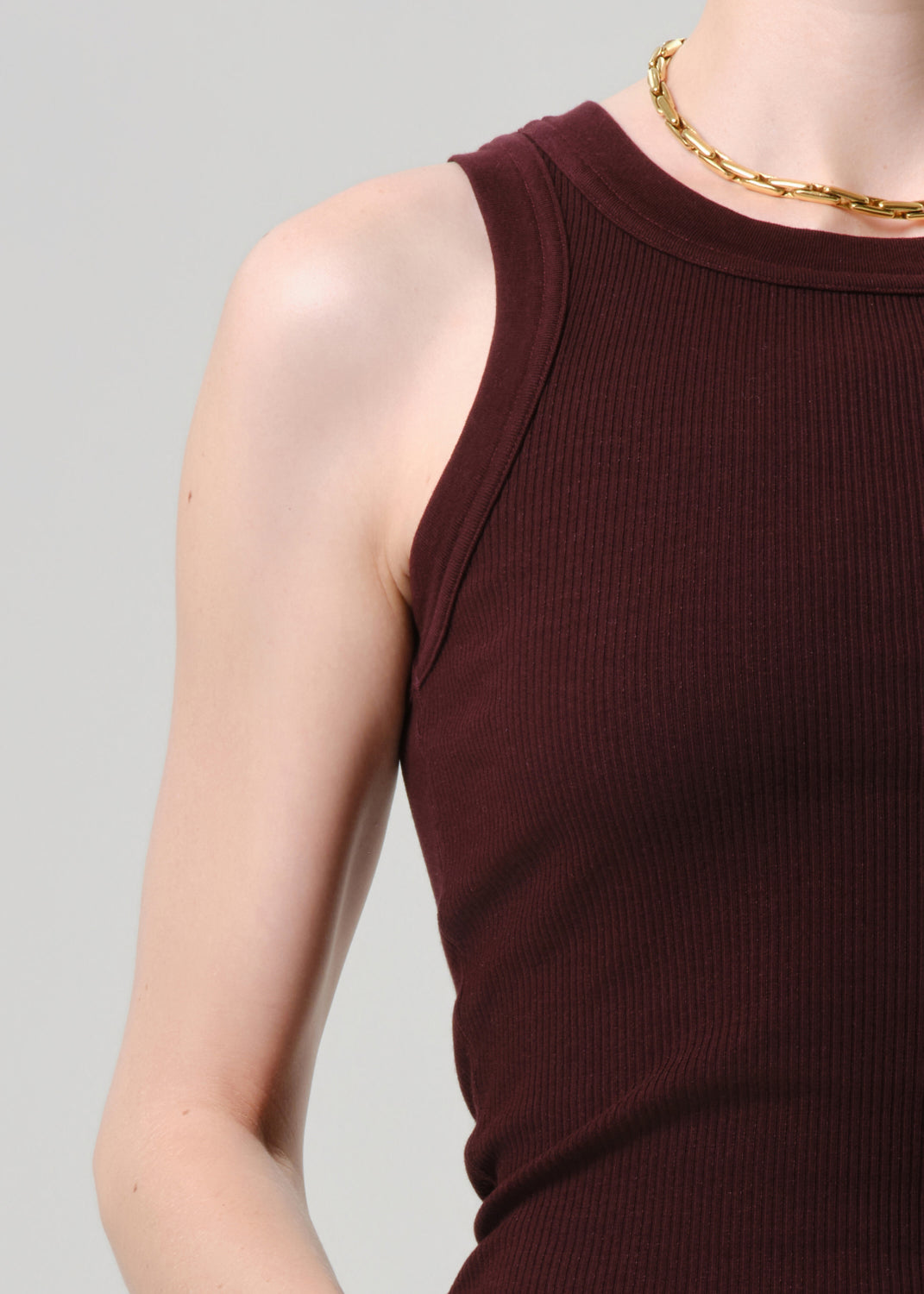 Close-up of a person wearing a ribbed sleeveless tank top in deep burgundy, paired with a chunky gold chain necklace, against a grey background.