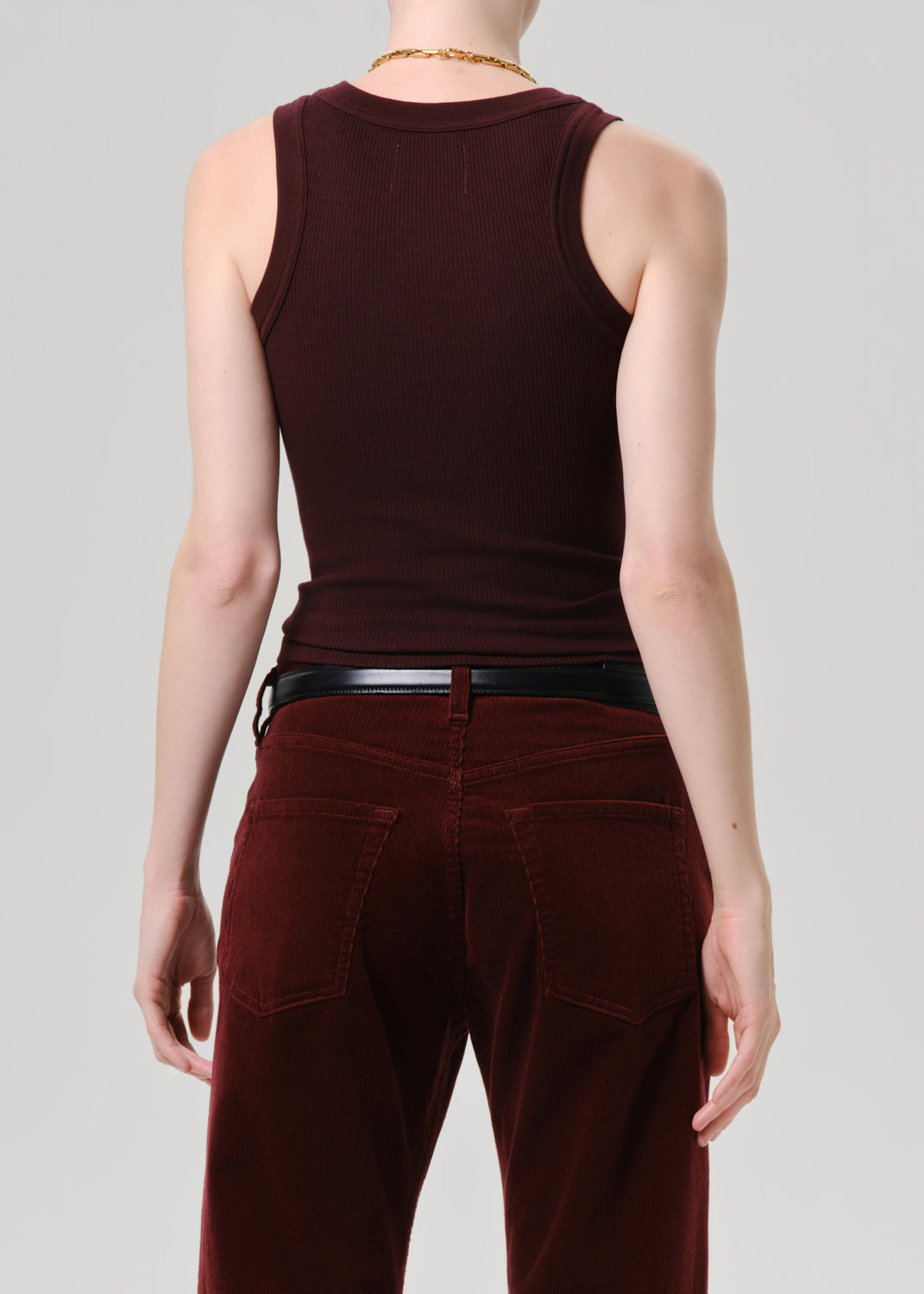 Rear view of a model wearing a dark burgundy ribbed tank top and matching burgundy corduroy pants with a black belt, against a grey background.