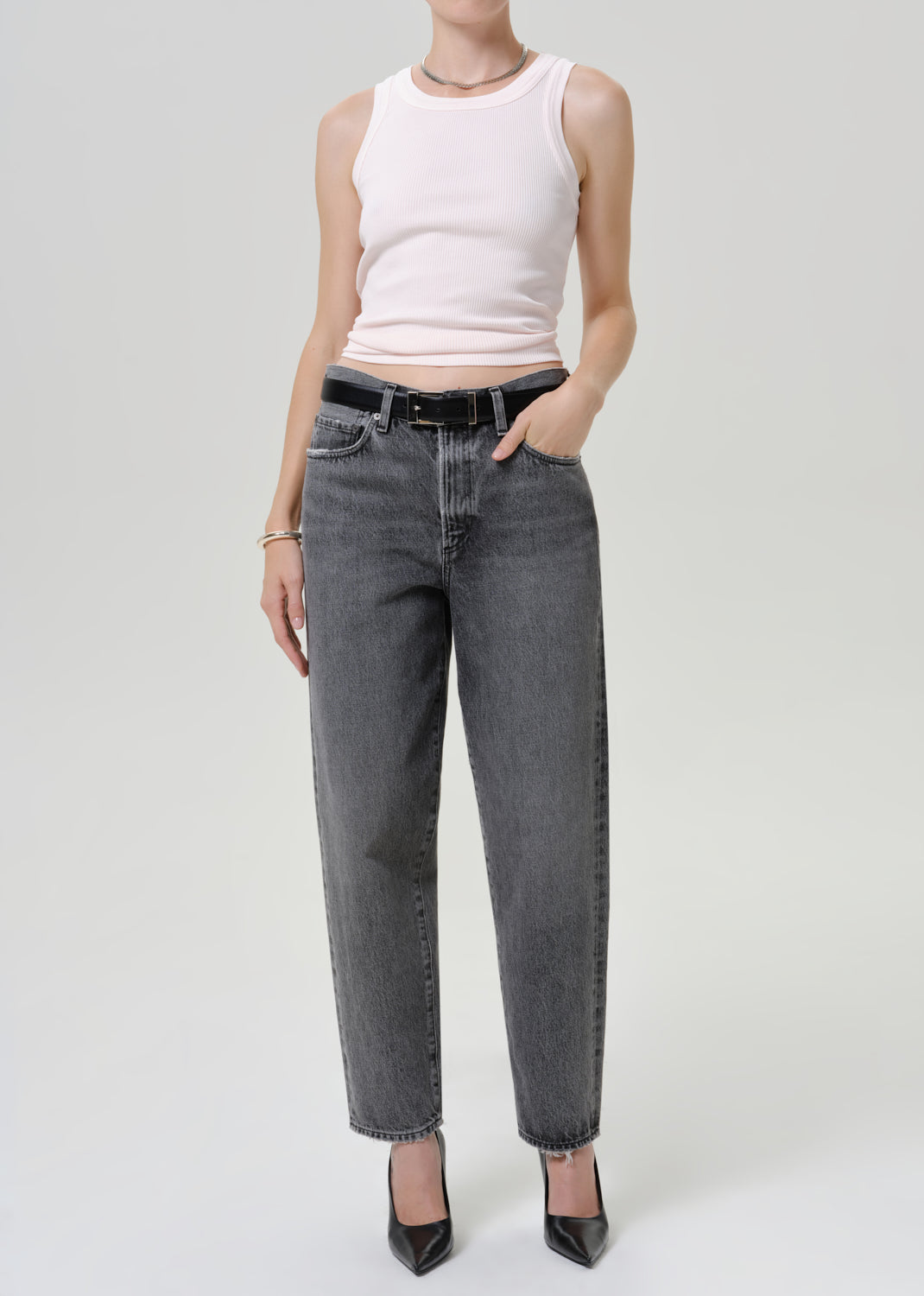 A model wearing a light pink ribbed tank top and dark gray straight-leg jeans with a black belt, paired with black pointed-toe heels, against a light gray studio background.
