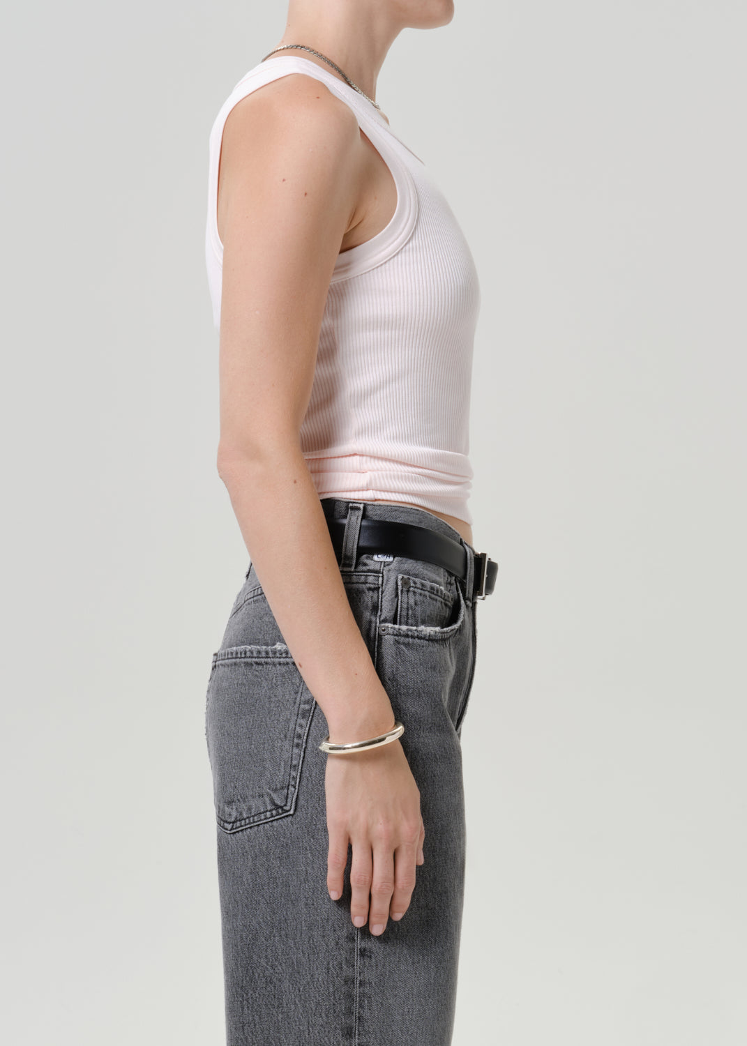 Side profile of a model wearing a light pink ribbed tank top, gray jeans, a black belt, and a silver bracelet against a light gray background.