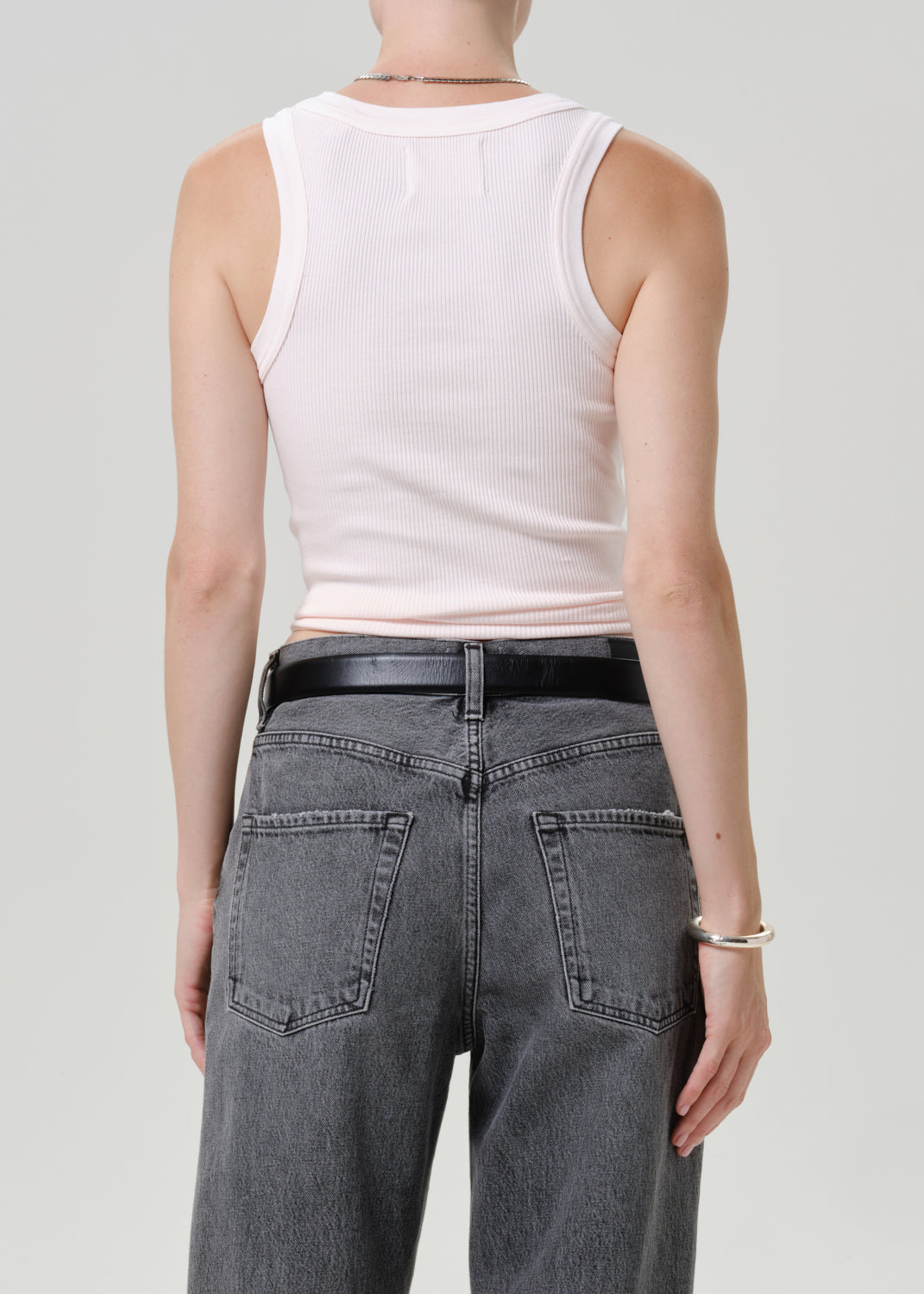 Rear view of a model wearing a pale pink ribbed tank top and gray jeans with a black belt, against a light gray studio background.