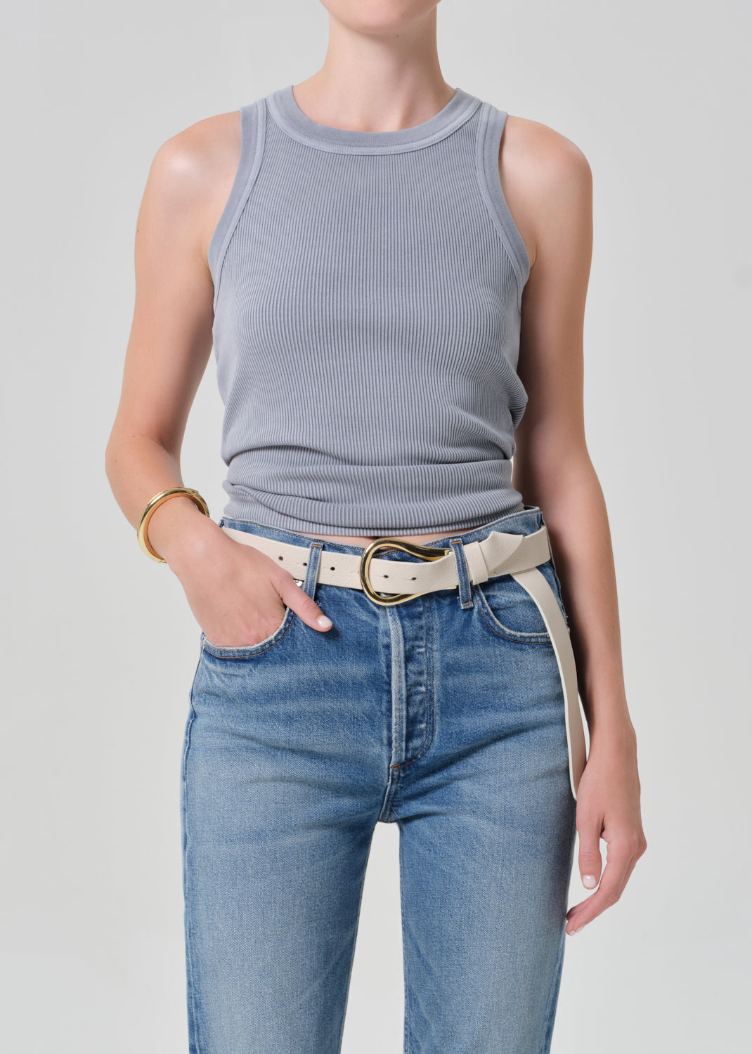 Person wearing a gray tank top and blue jeans with a belt on a plain background