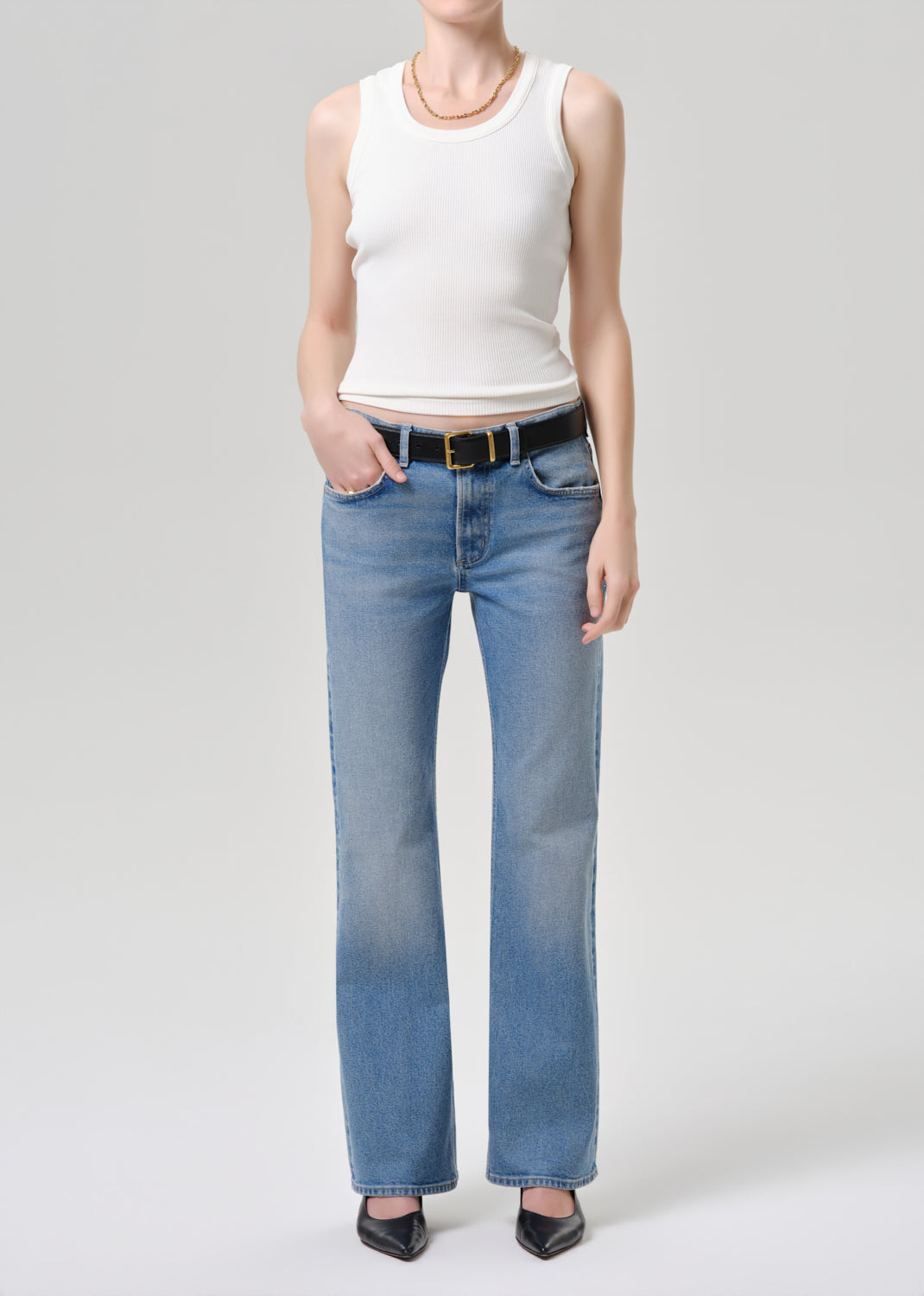 A model wearing light-wash blue straight-leg jeans with a black belt, paired with a white ribbed tank top and black pointed-toe heels, photographed against a grey background.
