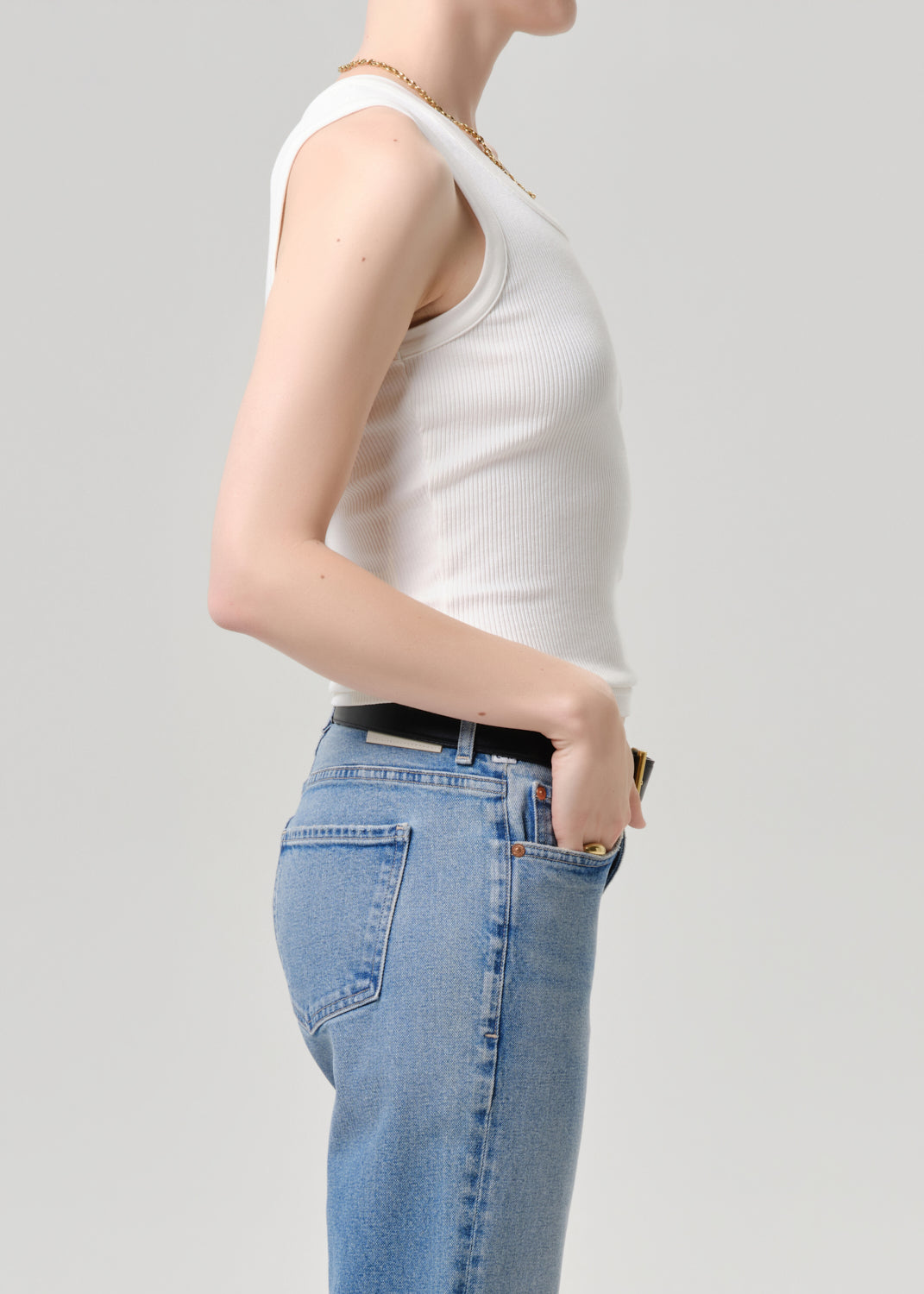 A side-profile view of a model wearing a white ribbed tank top and light blue jeans with a black belt, set against a grey background.
