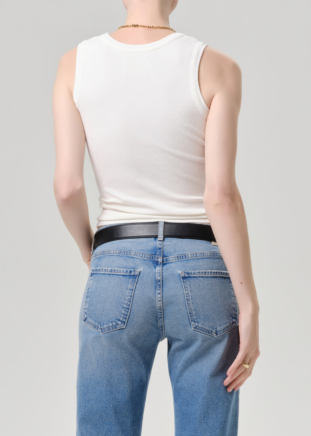 Rear view of a model wearing a white ribbed tank top, a black leather belt, and light blue jeans against a grey background.