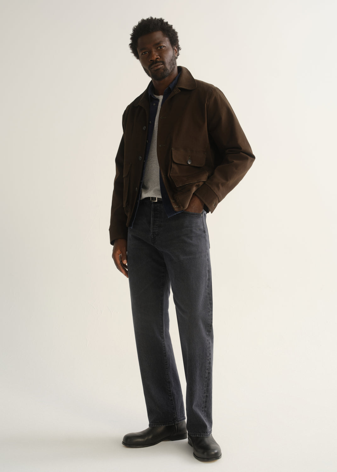Person wearing washed charcoal grey straight leg jeans with a dark brown jacket, dark shirt, and black shoes, angled front full body view on a light grey background.