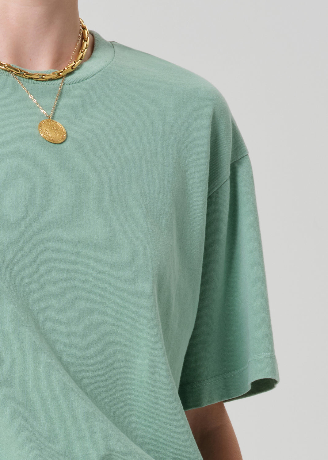 Person wearing a light green t-shirt and gold necklaces on a plain background