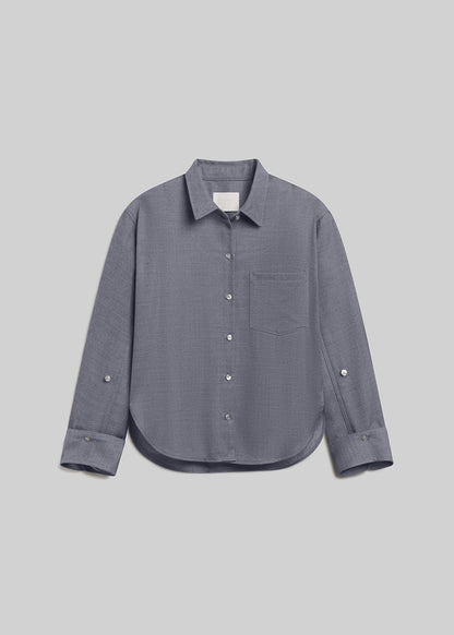 Gris grey long-sleeve wool button-down shirt with a pointed collar, front button closure, single chest pocket, buttoned cuffs, and curved hem, front flat lay view on a grey background.
